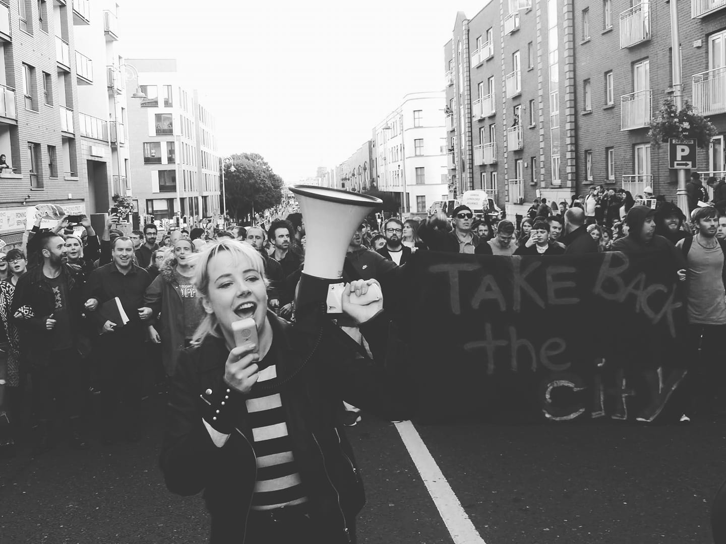 Protesters of the Take Back the City Dublin movement are walking down the street. A girl is holding a megaphone. The activists militate for a right to housing.