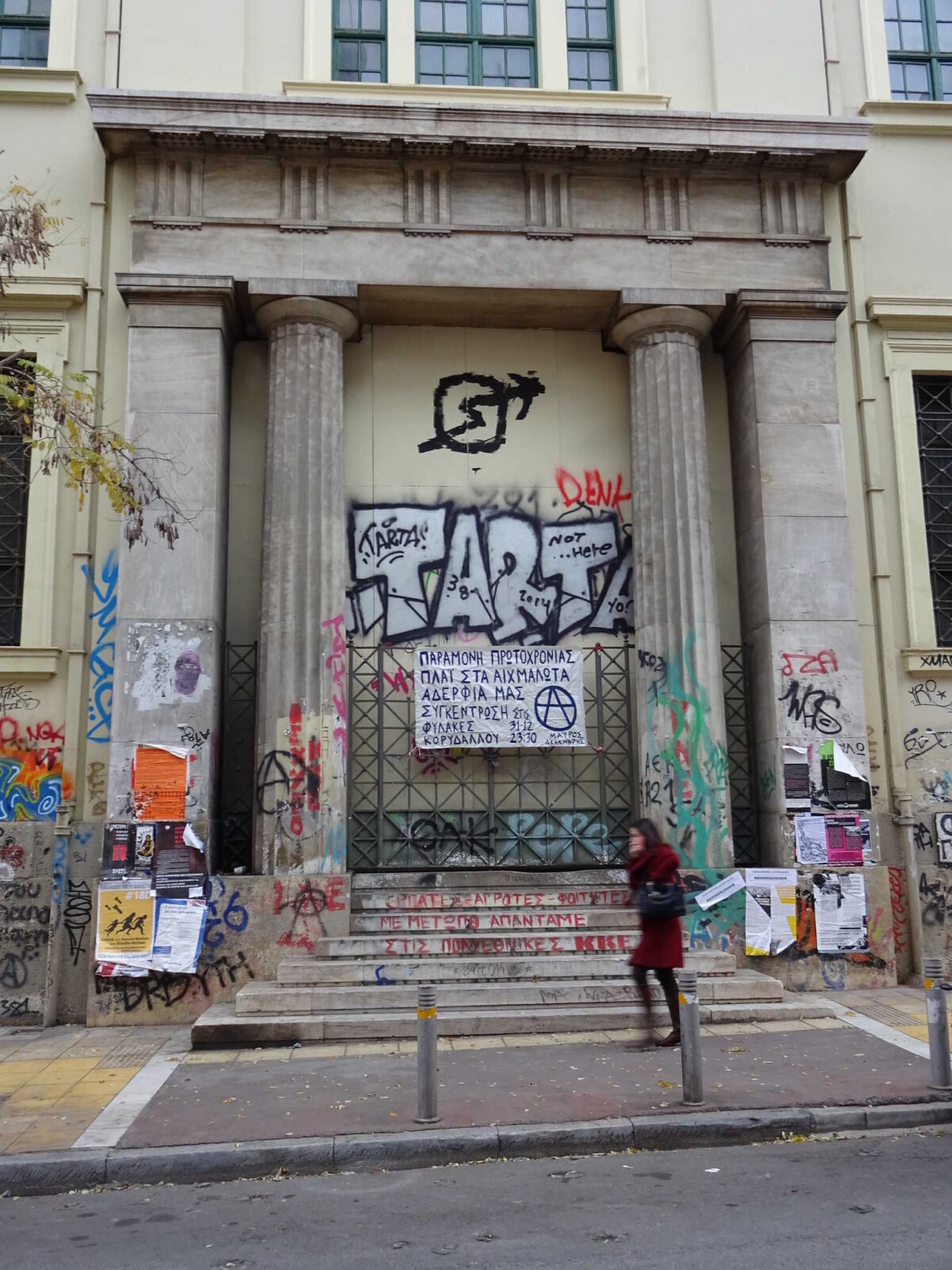 Image of a front door of the polytechnical Univserity in Athen, Exarchia. On the upper side oyu can see the sign of the political squat movement: A circle with an arrow crossing trough. A person is walking by the steps leading to the door.