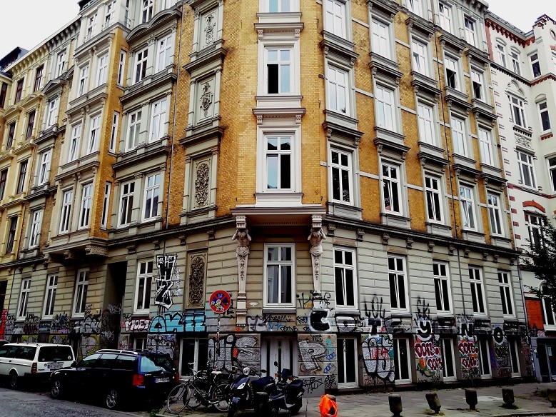 The facade of an building in Berlin made of yellow bricks looks like built around 1920. In front of the building are some cars, bikes and scooters. The walls are full of different Graffiti. Image by Urbanauth.