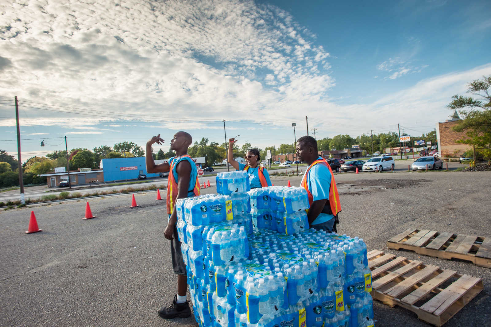 Trinkwasserflaschen auf Paletten mit Helfern in orangenen Jacken auf einem Parkplatz in Flint, Michigan, USA. Flint's Wasserkrise führte dazu, dass das Wasser aus dem Wasserhahn nicht mehr bekömmlich war. Himmel blau leicht bewölkt. Helfer sind people of color.
