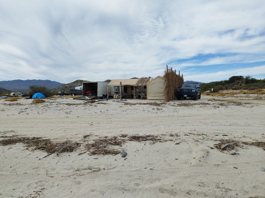 Van-Life in einer Hochwasser Risikozone in San Bartolo, Baja California Sur, Mexiko. Auf dem Sand steht ein Wohnmobil mit Terrasse, Container und improvisierten Parkplatz.  